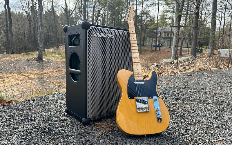 Black Soundboks 4 outdoors speaker with an orange Fender guitar on a gravel patio