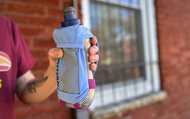 A person holding a Nathan QuickSqueeze Insulated Water bottle against a brick wall.
